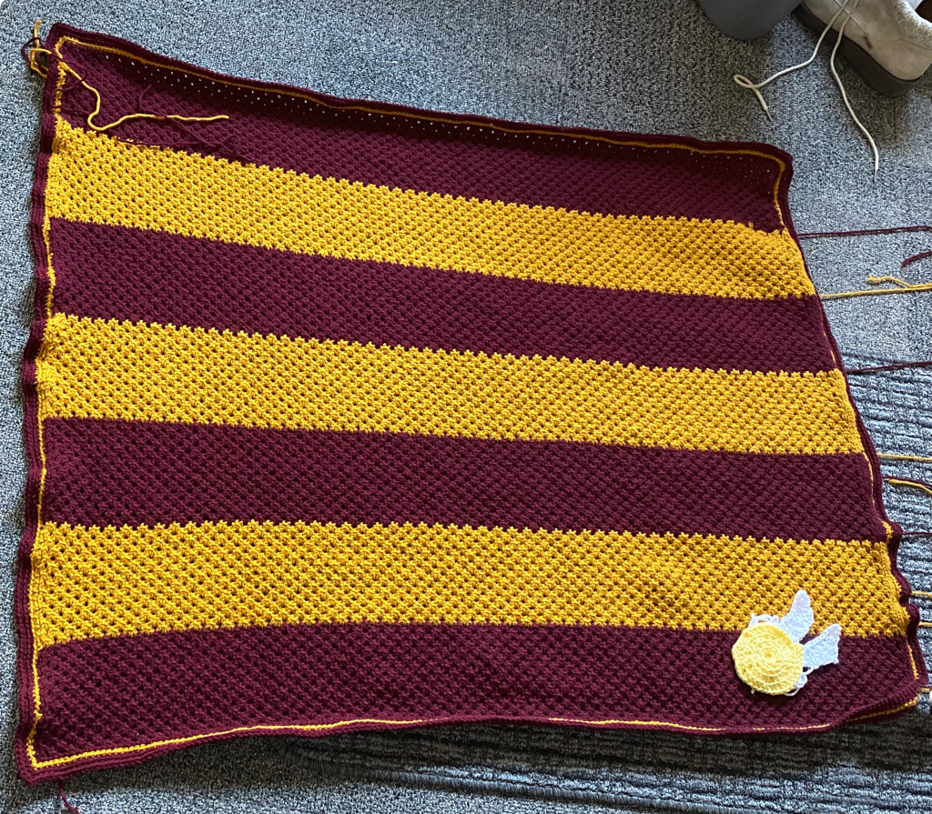 Another image of the striped baby blanket, the stripes run horizontally now a golden snitch has been added in the bottom corner with two white wings stacked above one another